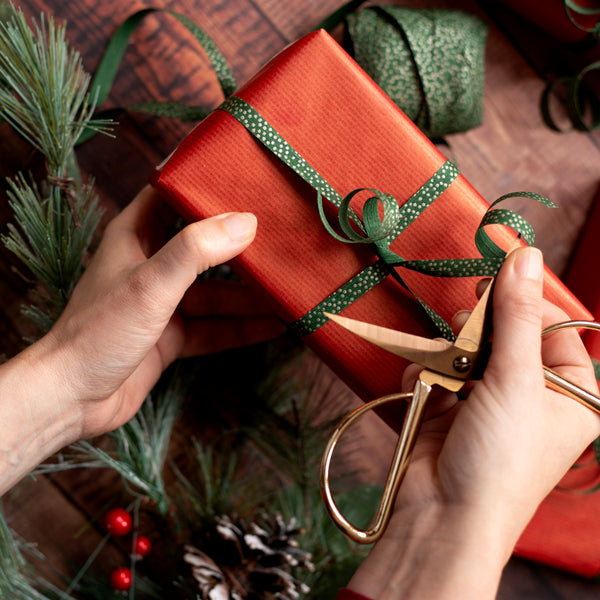 Person curling green ribbon on a red gift box using gold scissors, surrounded by Christmas decorations.