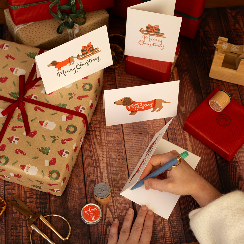 Person writing a card next to wrapped gifts on a wooden surface