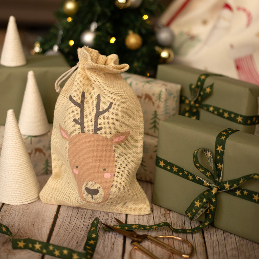 Christmas-themed gift bag with reindeer design on a wooden surface with wrapped presents and a tree in the background.