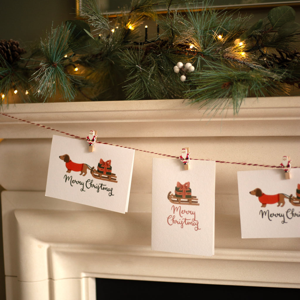 Christmas cards with sausage dog and sleigh designs hanging on a fireplace mantle.