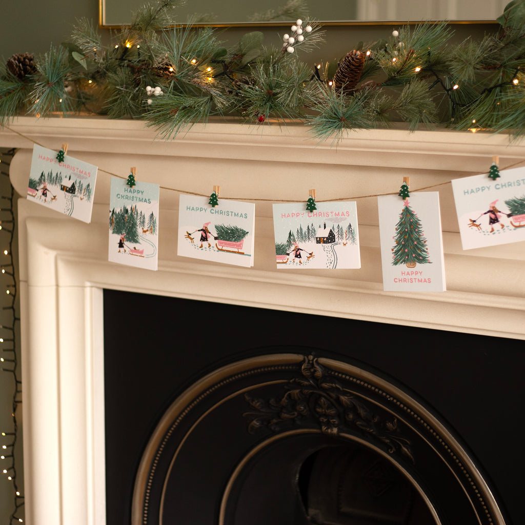 Christmas cards on a string with clips against a fireplace mantel decorated with greenery and lights.