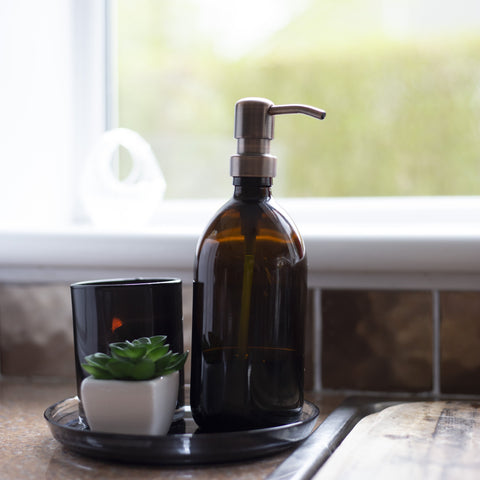 refillable soap bottle with brass pump head sits by a sink in front of a window