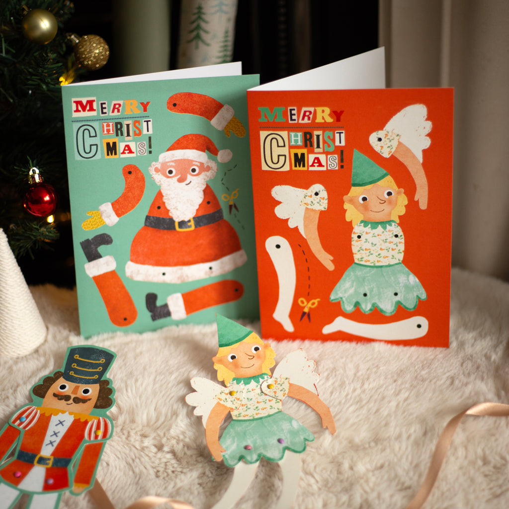 Two Christmas cards with festive illustrations of Santa Claus and a fairy, placed on a textured surface with a Christmas tree in the background.