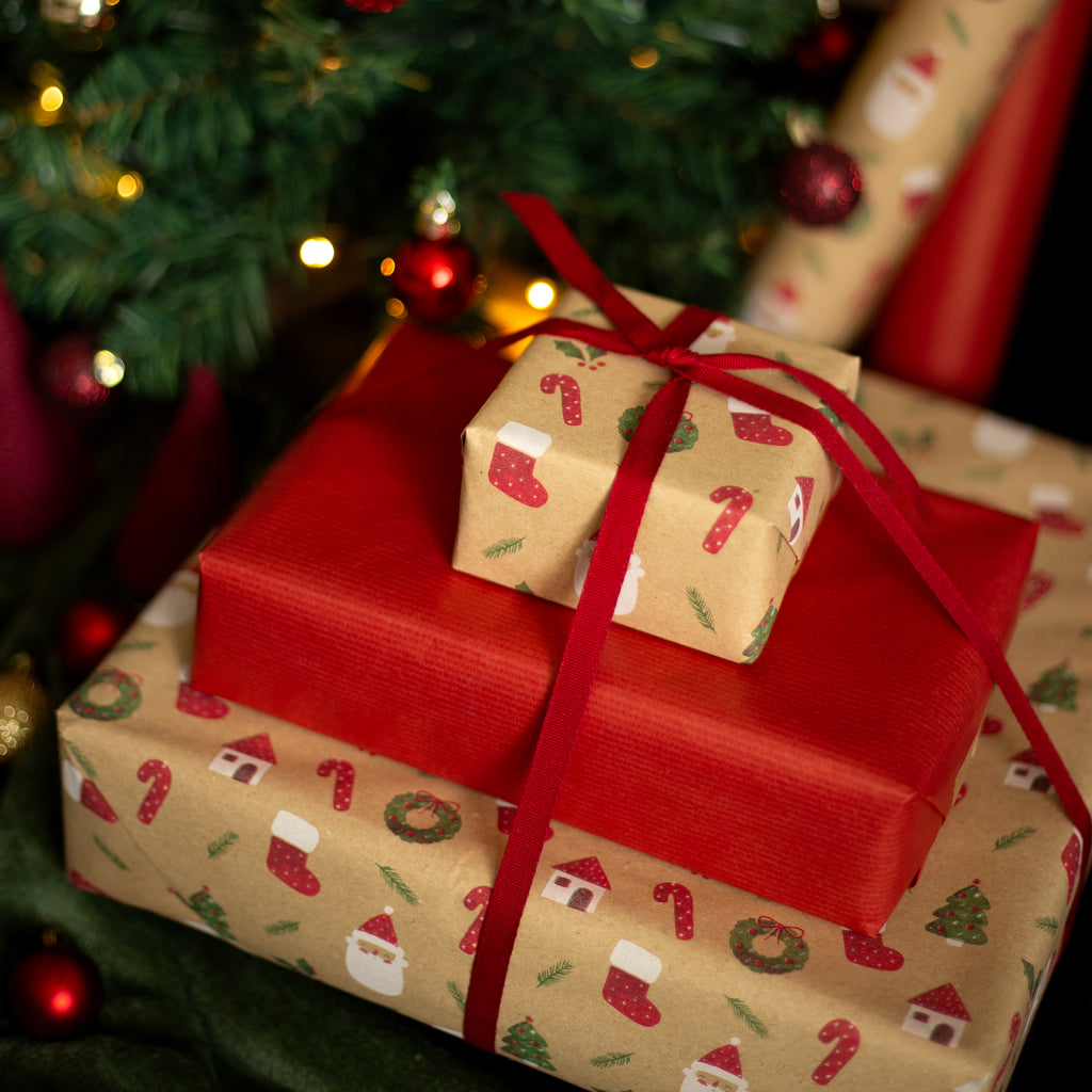 Stack of Christmas presents wrapped in red and brown paper with festive designs, tied with red ribbons, against a blurred Christmas tree background.