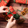 Person wrapping a small red gift box with green ribbon on a wooden surface with decorative elements.