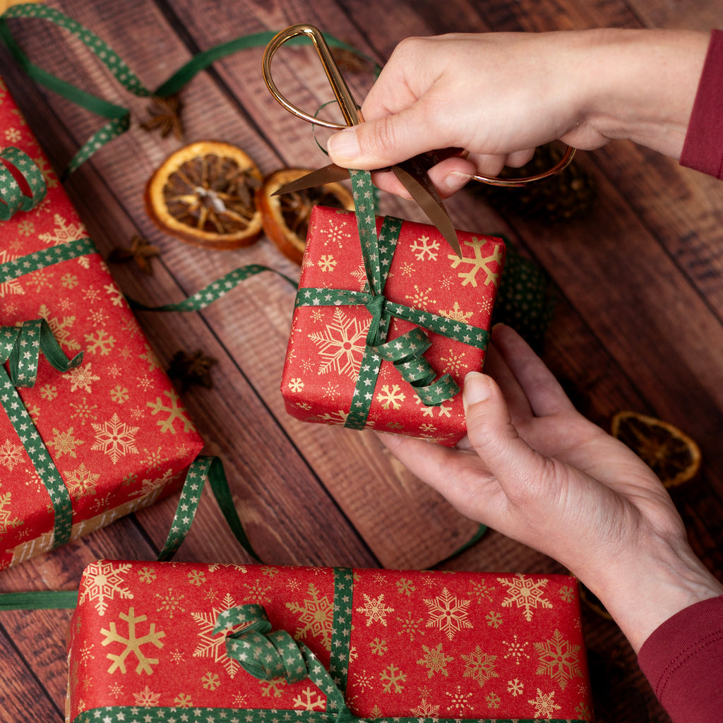 Person wrapping a small red gift box with green ribbon on a wooden surface.