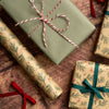 Christmas-themed gift wrapping set featuring beige wrapping paper with a green pine branch pattern and red ribbon accents, alongside solid sage green wrapping paper, arranged on a rustic wooden surface with pine branches and red berries.