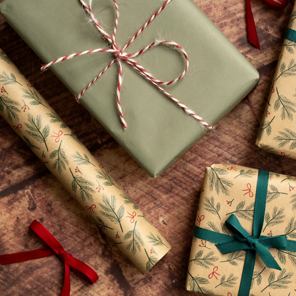 Christmas-themed gift wrapping set featuring beige wrapping paper with a green pine branch pattern and red ribbon accents, alongside solid sage green wrapping paper, arranged on a rustic wooden surface with pine branches and red berries.