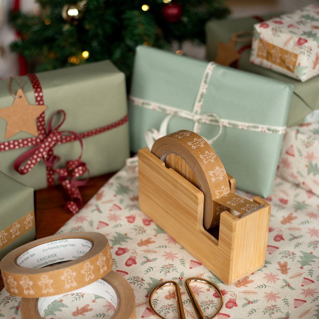 Wooden tape dispenser with decorative tape and scissors on a festive wrapping paper with Christmas presents in the background.