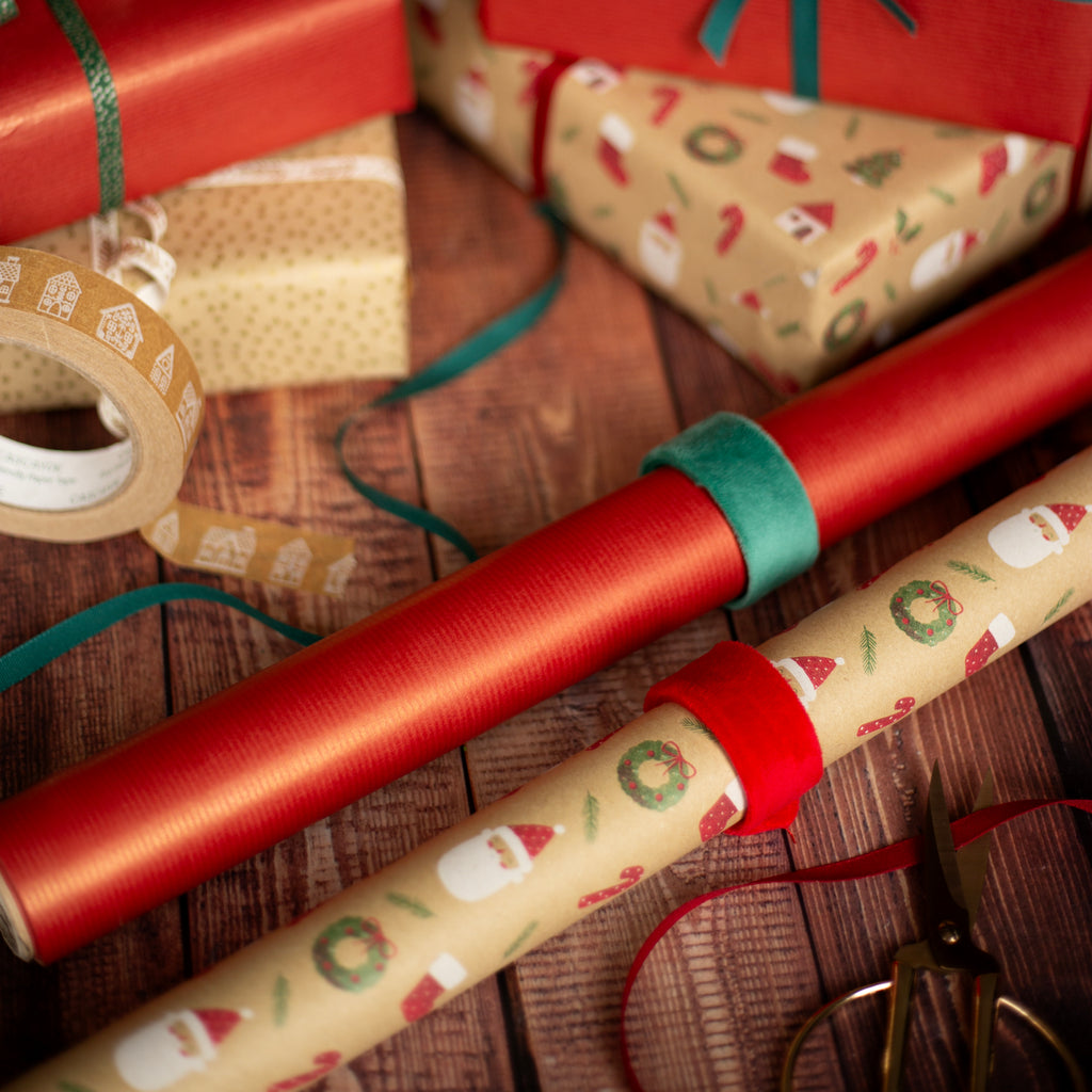 Rolls of Christmas-themed wrapping paper with sealing strips on a wooden surface with tape and scissors.