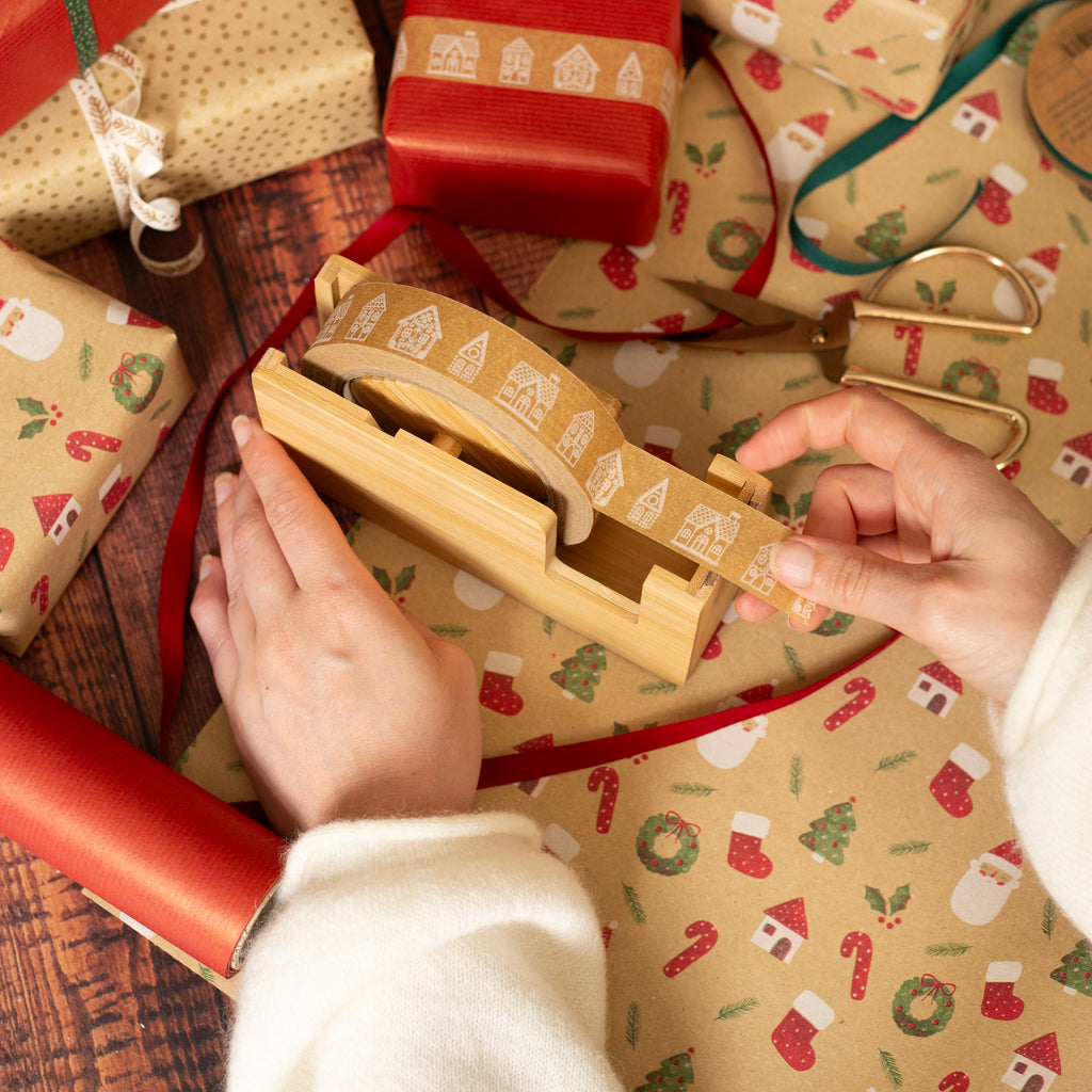 Person pulling Gingerbread House paper tape from a bamboo dispenser with Christmas-themed wrapping paper in the background