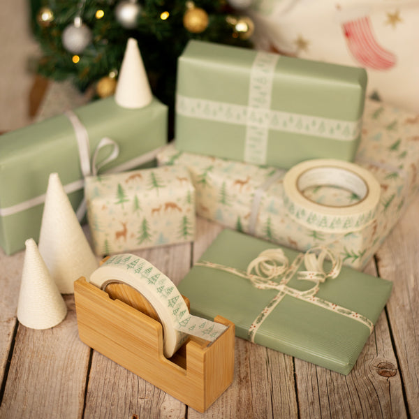 Green gift boxes with green fir tree tape and ribbon on a wooden surface with a Christmas tree in the background.