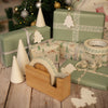 Christmas gift wrapping with green boxes, decorative tape, and tree-shaped cones on a wooden surface.