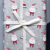 Silver wrapping paper with a repeating Santa Claus and snowman pattern, shown both as a rolled sheet and wrapped around a gift box tied with a white ribbon on a split light blue and dark navy background.