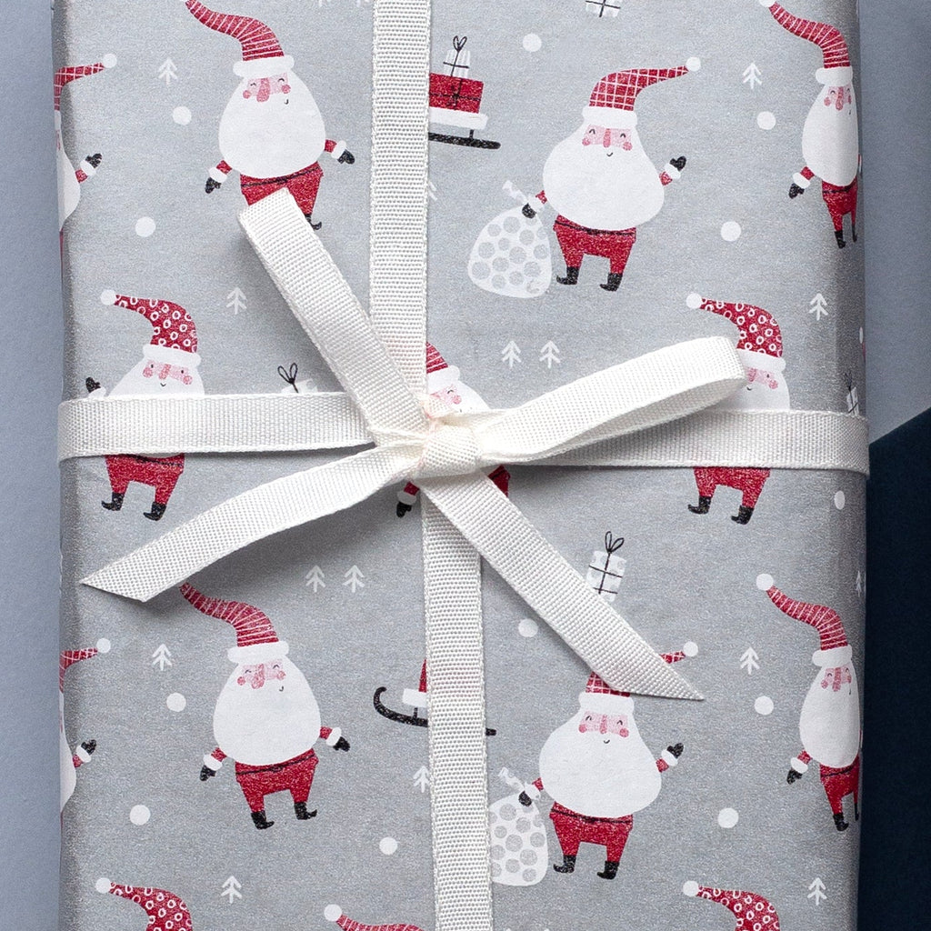 Silver wrapping paper with a repeating Santa Claus and snowman pattern, shown both as a rolled sheet and wrapped around a gift box tied with a white ribbon on a split light blue and dark navy background.
