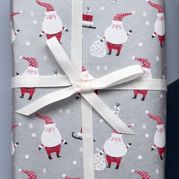 Silver wrapping paper with a repeating Santa Claus and snowman pattern, shown both as a rolled sheet and wrapped around a gift box tied with a white ribbon on a split light blue and dark navy background.