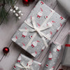 Three wrapped Christmas presents arranged on a whitewashed wooden surface with pine branches, red ornaments, and white berries. The center gift is wrapped in light gray paper with a gnomes print and tied with a white ribbon, while the other two gifts are wrapped in metallic silver paper with red ribbon and bow accents.