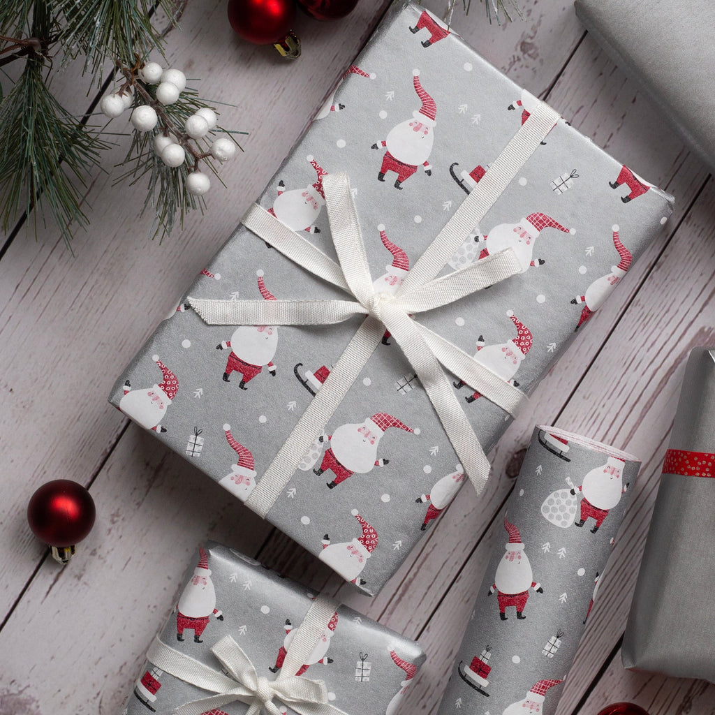Three wrapped Christmas presents arranged on a whitewashed wooden surface with pine branches, red ornaments, and white berries. The center gift is wrapped in light gray paper with a gnomes print and tied with a white ribbon, while the other two gifts are wrapped in metallic silver paper with red ribbon and bow accents.