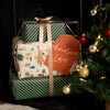 Three wrapped Christmas gifts stacked together with a beige ribbon and a red paper tag that reads 'Christmas Love'. The wrapping paper features green diagonal stripes and a festive holiday print, set next to a decorated Christmas tree.