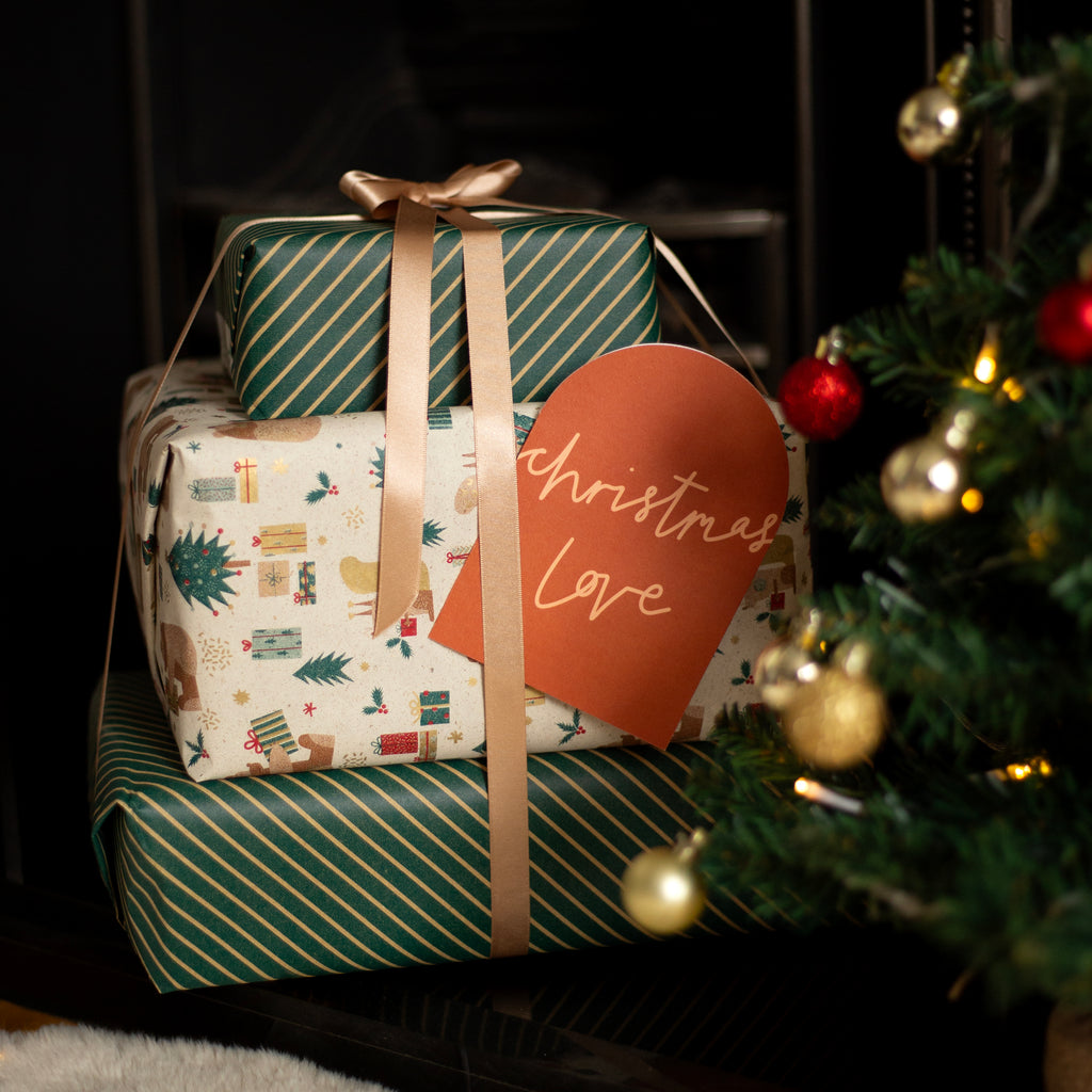 Three wrapped Christmas gifts stacked together with a beige ribbon and a red paper tag that reads 'Christmas Love'. The wrapping paper features green diagonal stripes and a festive holiday print, set next to a decorated Christmas tree.