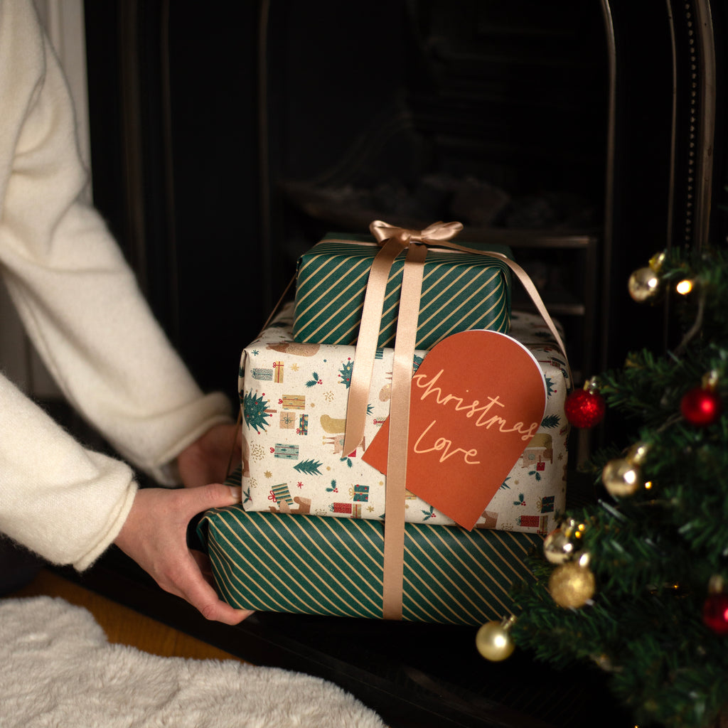Three stacked Christmas gift boxes wrapped in festive paper, held by a person wearing a white sweater near a decorated Christmas tree. The wrapping includes green striped paper, a cream paper with small holiday illustrations, and a brown paper tag that reads 'Christmas love'.