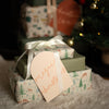 A stack of wrapped Christmas presents on a soft white faux-fur surface, with a blurred Christmas tree and ornaments in the background. The wrapping paper has a woodland deer and tree print in muted green, beige, and brown tones, with one box wrapped in solid sage green paper.