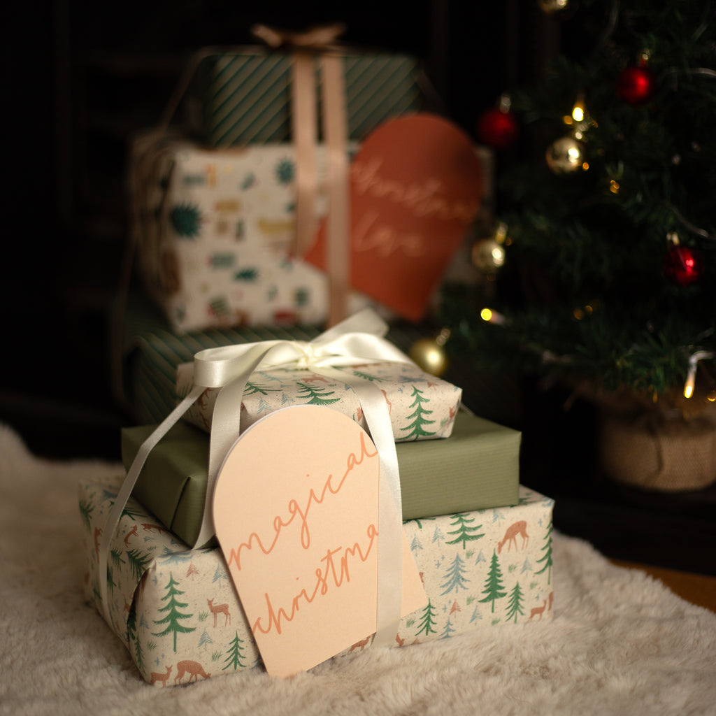 A stack of wrapped Christmas gift boxes on a white faux-fur surface, with a blurred Christmas tree in the background. The front box is wrapped in a light green and white forest-patterned paper with a cream ribbon and a peach tag that reads 'magical christmas'.