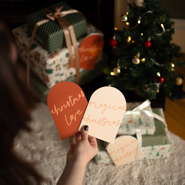 A person holds two Christmas-themed paper tags or labels in a cozy holiday setting. One tag is a redish orange with the words 'christmas love' in rose-gold script, and the other is cream with 'magical christmas' in rose-gold script.