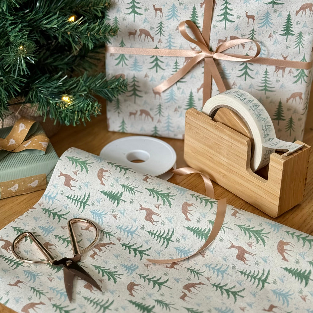 Christmas wrapping paper with forest design, ribbon, tape, and scissors on a wooden surface.