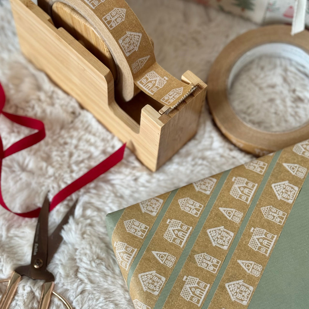 Wooden tape dispenser with patterned tape, scissors, and red ribbon on a textured surface.