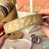 Kraft Brown Paper tape with white floral & Fern patterns held by a hand, with a wrapped gift and ribbons and stamps in the background.