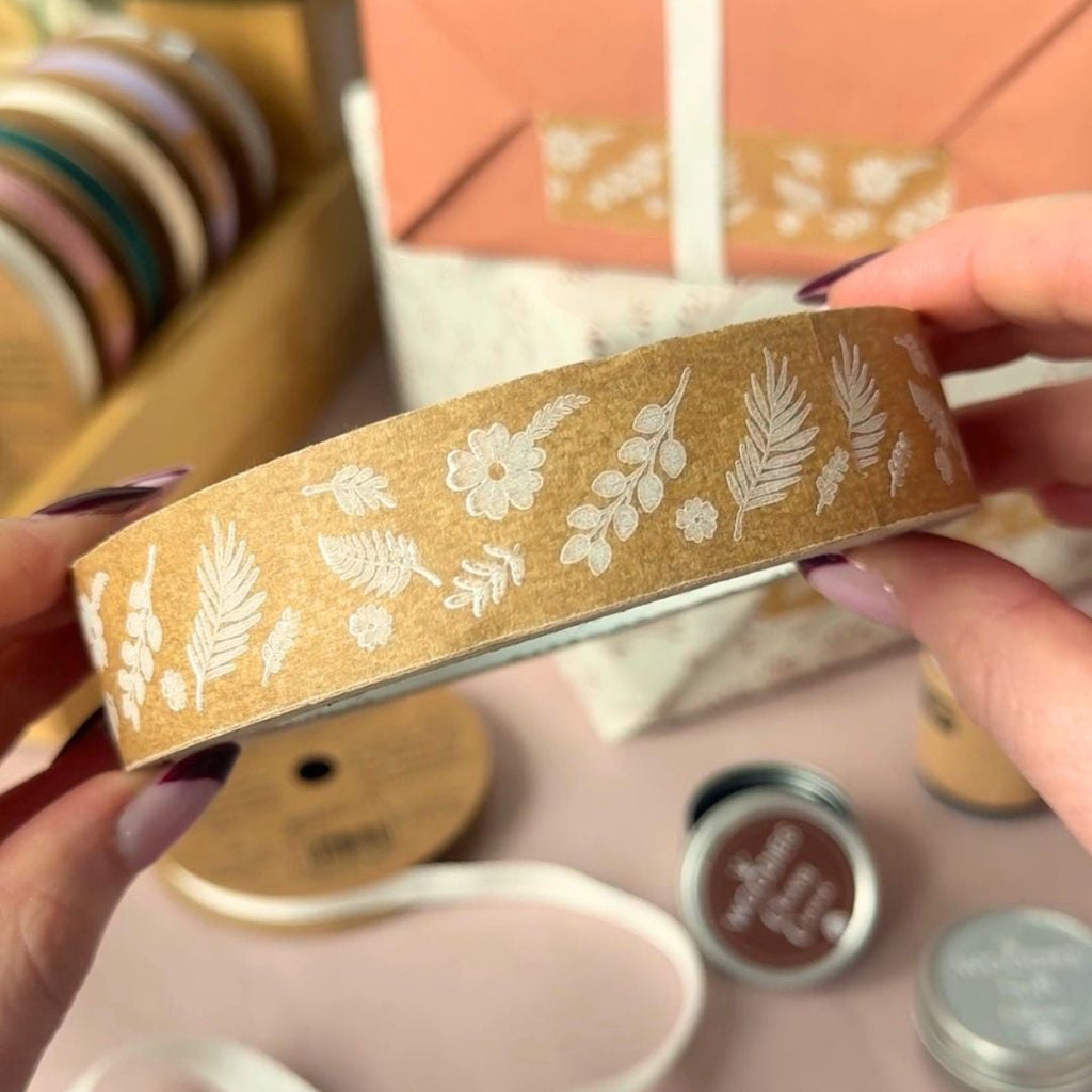 Kraft Brown Paper tape with white floral & Fern patterns held by a hand, with a wrapped gift and ribbons and stamps in the background.