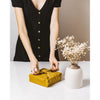 a tall lady in a black dress wraps a gift in yellow Furoshiki material gift wrap next to a vase of dried white flowers