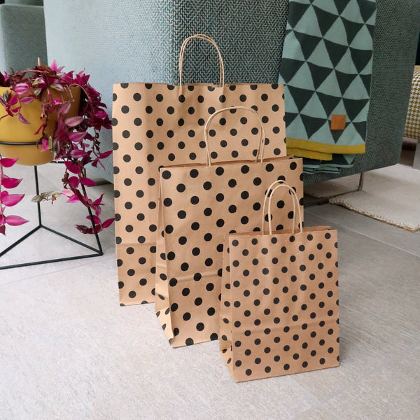 3 brown paper gift bag with black polka dots sits on the floor next to a green material sofa and through and pink house plant
