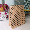 brown paper gift bag with black polka dots sits on the floor next to a green material sofa and through and pink house plant