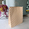 brown paper gift bag with white polka dots sits on the floor next to a green material sofa and through and pink house plant