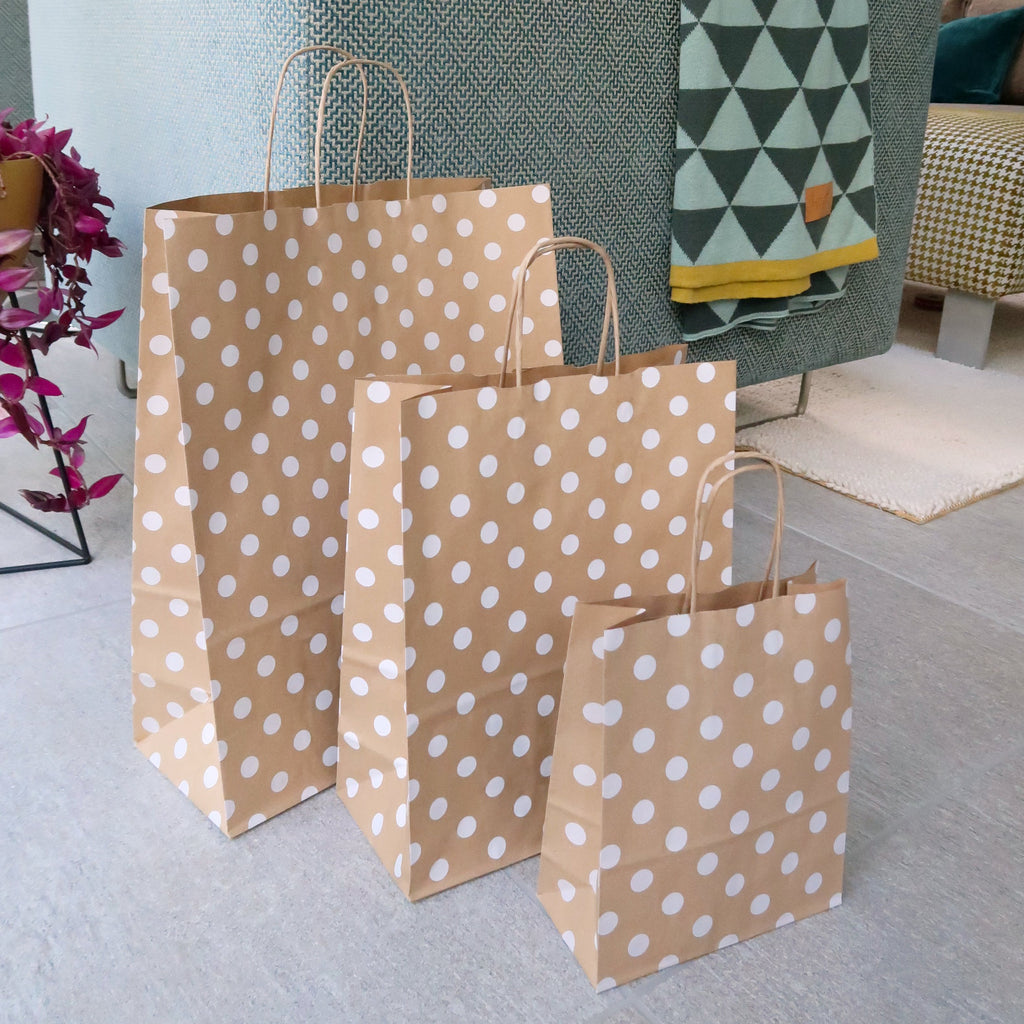 3 brown paper gift bag with white polka dots sits on the floor next to a green material sofa and through and pink house plant